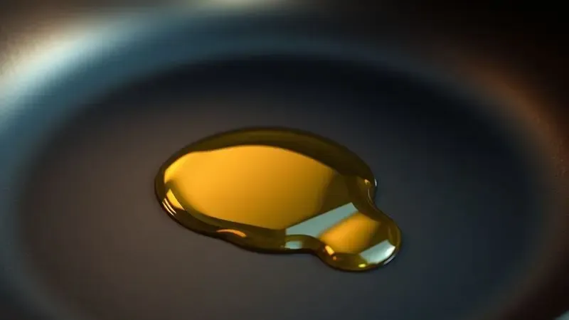 Ilustração 3D detalhando uma gota de óleo sendo espalhada uniformemente sobre a superfície escura de uma frigideira para criar uma camada protetora.